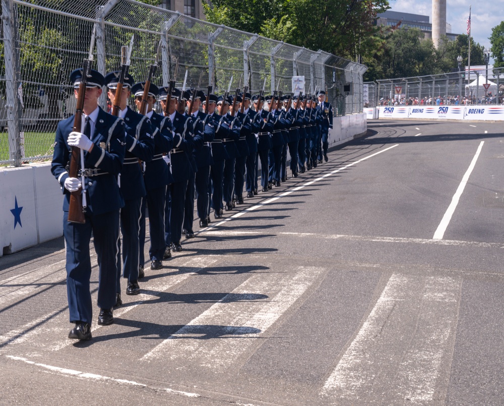 A precision drill team in dark blue military dress uniforms marches in a tight single-file formation along a paved circuit or street course. The personnel are carrying rifles and wearing white gloves and service caps, consistent with a ceremonial honor guard performance. Sponsor barriers bearing 'Boeing' and '250' branding line the course, with spectators visible in the background.