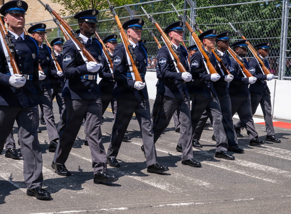 A precision drill team in dark navy blue dress uniforms is marching in formation during a formal military parade or ceremony. The personnel are carrying rifles at a uniform angle and wearing matching caps with blue bands, white gloves, and polished black shoes. The formation appears highly disciplined and synchronized, suggesting a ceremonial honor guard unit.