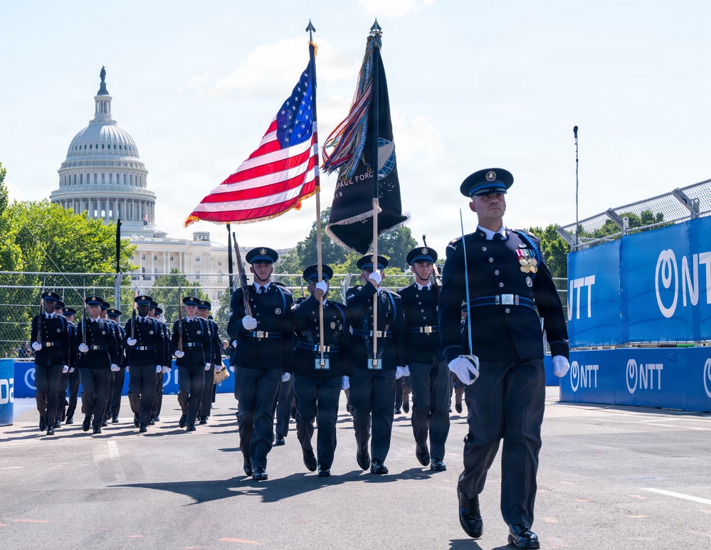 A U.S. Space Force honor guard is marching in a formal parade near the U.S. Capitol building in Washington, D.C. The formation carries the American flag and a black U.S. Space Force guidon flag, led by an officer in dress uniform with medals. The event appears to be associated with an NTT-sponsored venue or race circuit, based on the prominent blue barriers lining the route.