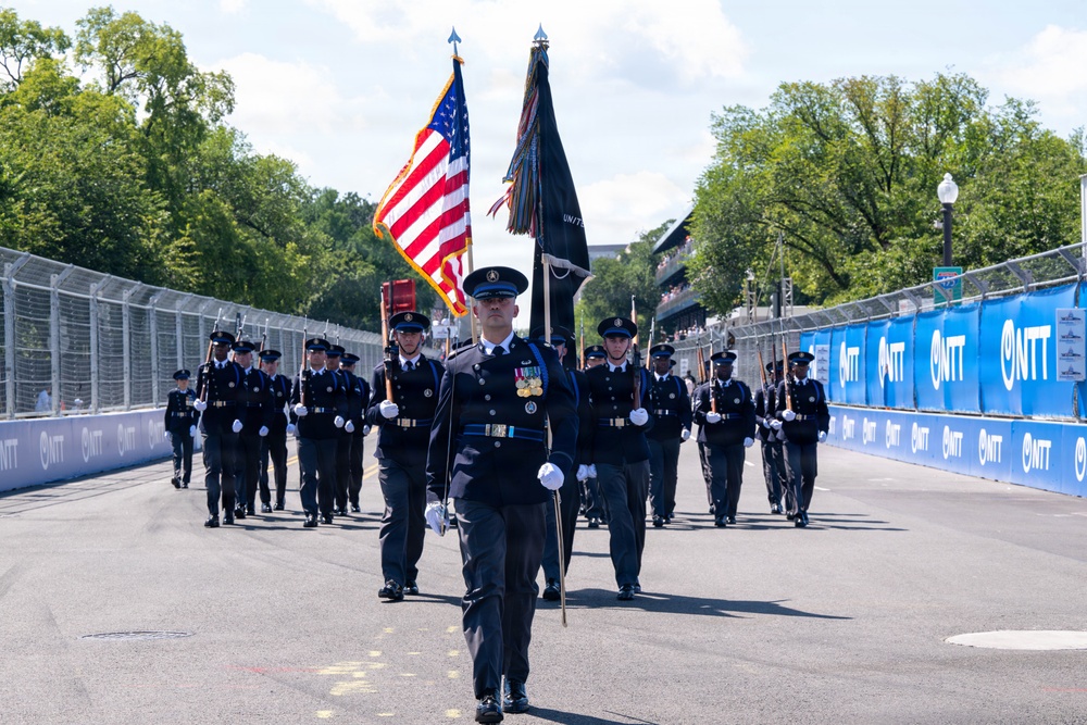 A military or law enforcement honor guard is marching in formation down a racing circuit or street course. The procession features flag bearers carrying the American flag and a dark unit flag, with personnel in formal dark uniforms and white gloves. The event appears to be a pre-race ceremony at a motorsport event, evidenced by the NTT-branded barriers lining the track.