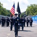 A military or law enforcement honor guard is marching in formation down a racing circuit or street course. The procession features flag bearers carrying the American flag and a dark unit flag, with personnel in formal dark uniforms and white gloves. The event appears to be a pre-race ceremony at a motorsport event, evidenced by the NTT-branded barriers lining the track.