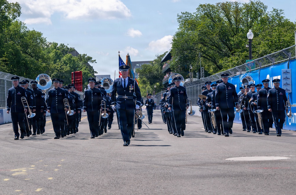 A military marching band in dark dress uniforms parades down a closed street lined with blue barriers and signage. The formation is led by an officer carrying a ceremonial sword, flanked by color guard members bearing flags including the American flag. The setting appears to be an outdoor event venue or racing circuit, with trees and buildings visible in the background.