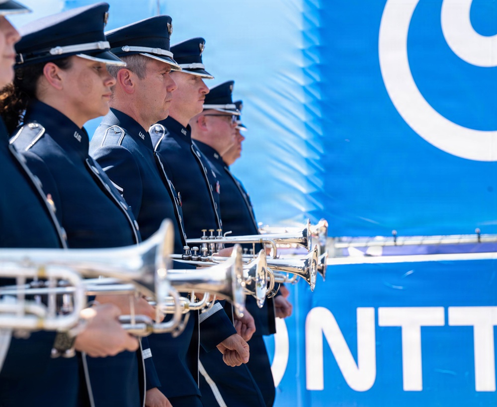 A group of uniformed military band members stand in a line holding silver trumpets/bugles at attention. They are wearing dark navy dress uniforms with peaked caps bearing silver insignia, consistent with U.S. Air Force ceremonial dress. The scene appears to be an outdoor official event, with a blue banner bearing partial white text 'NTT' visible in the background.