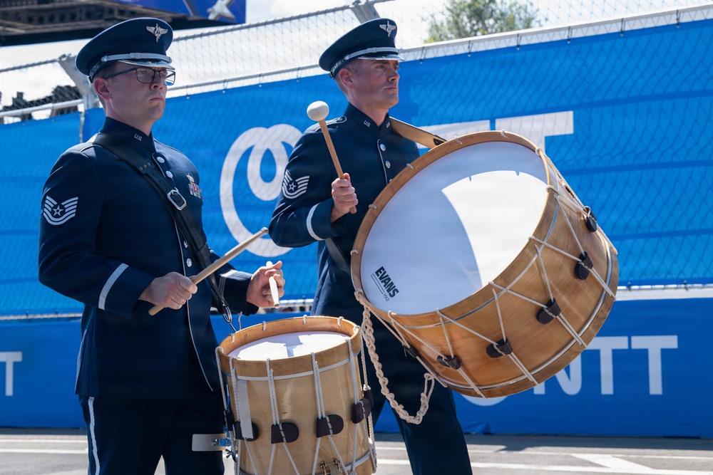 Two U.S. Air Force military band members in dress uniforms are playing drums at an outdoor event. Both wear Technical Sergeant rank insignia and are positioned in front of blue sponsor banners. The drummer on the right is playing a large bass drum with an Evans drumhead, while the one on the left plays a smaller field drum.