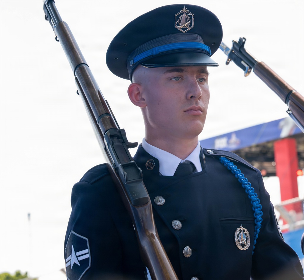 A young military service member in a formal dress uniform stands at attention holding a rifle, appearing to participate in a ceremonial guard or honor guard event. The uniform features distinctive Space Force insignia and a blue aiguillette, indicating this is a U.S. Space Force ceremonial unit. The subject maintains a serious, forward-focused expression consistent with formal military ceremony.