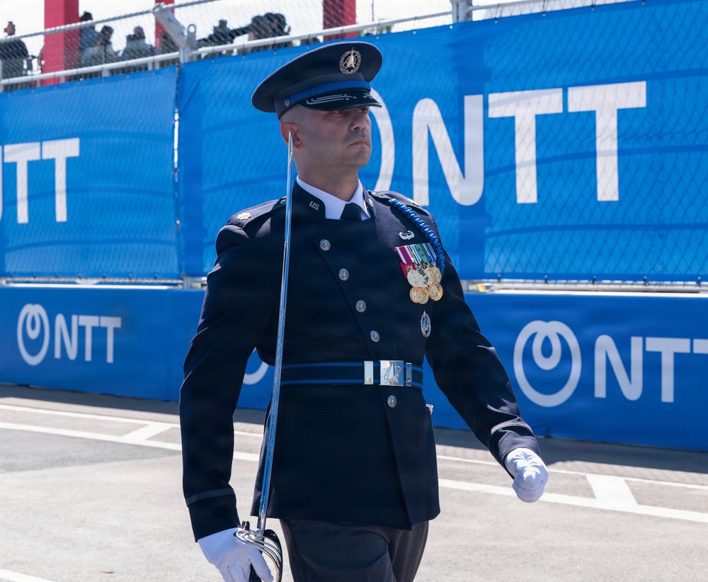 A military service member in formal dress uniform is marching or standing at attention at what appears to be a public event or ceremony. The individual is wearing a dark navy ceremonial uniform adorned with multiple medals, a peaked cap, white gloves, and is carrying a ceremonial sword. The backdrop features prominent blue NTT sponsor banners, suggesting this ceremony is taking place at a motorsport or large public venue.