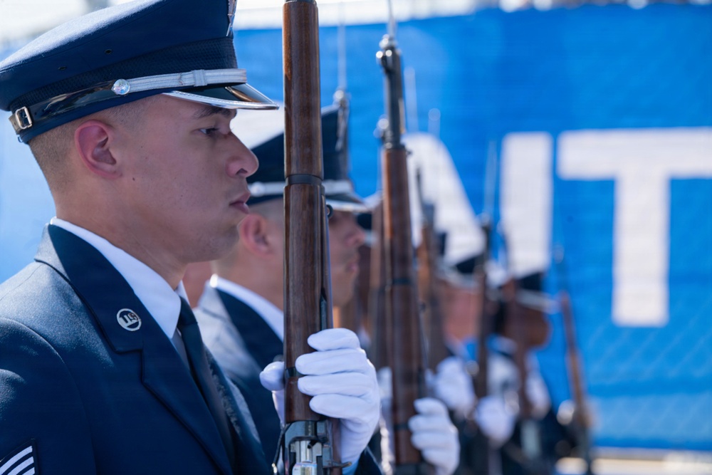 A line of U.S. Air Force Honor Guard members stand at attention in formal dress uniforms, each holding a rifle with wooden stock. The foreground airman is in sharp focus, wearing a dark blue service dress uniform with white gloves and a service cap. The formation extends into the background with a blue banner partially visible bearing white letters.