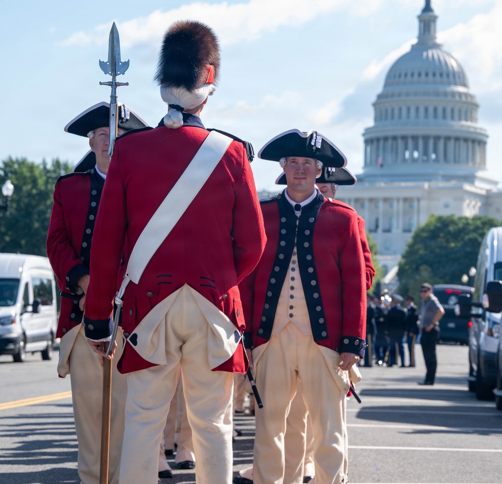 A group of performers or ceremonial soldiers dressed in 18th-century American Revolutionary War-era uniforms are assembled on a street in Washington, D.C. They wear red coats, tricorn hats, cream-colored breeches, and one figure prominently displays a tall bearskin hat and carries a halberd. The United States Capitol building is clearly visible in the background, suggesting this is a formal ceremonial or commemorative event.