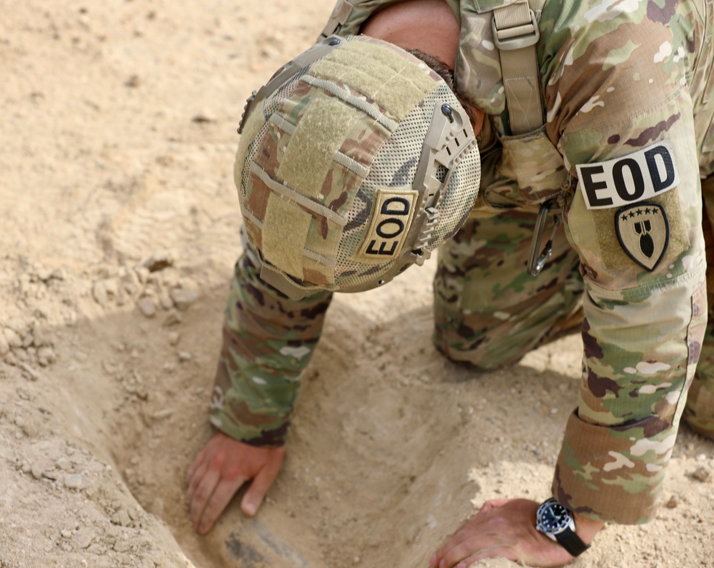 An EOD (Explosive Ordnance Disposal) technician in MultiCam/OCP camouflage uniform is kneeling in sandy terrain, examining or uncovering an object partially buried in the ground. The individual wears an EOD-marked helmet and has an EOD unit patch on the right sleeve. The scene suggests a field operation involving the identification or handling of a buried object.