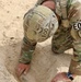 An EOD (Explosive Ordnance Disposal) technician in MultiCam/OCP camouflage uniform is kneeling in sandy terrain, examining or uncovering an object partially buried in the ground. The individual wears an EOD-marked helmet and has an EOD unit patch on the right sleeve. The scene suggests a field operation involving the identification or handling of a buried object.