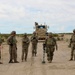 A group of approximately eight U.S. Army soldiers in MultiCam/OCP camouflage uniforms are gathered on a dirt road in an arid desert environment. One soldier is operating what appears to be a mine detector or metal detector device on the road surface. A tan MRAP (Mine-Resistant Ambush Protected) vehicle is positioned in the background on the same road.