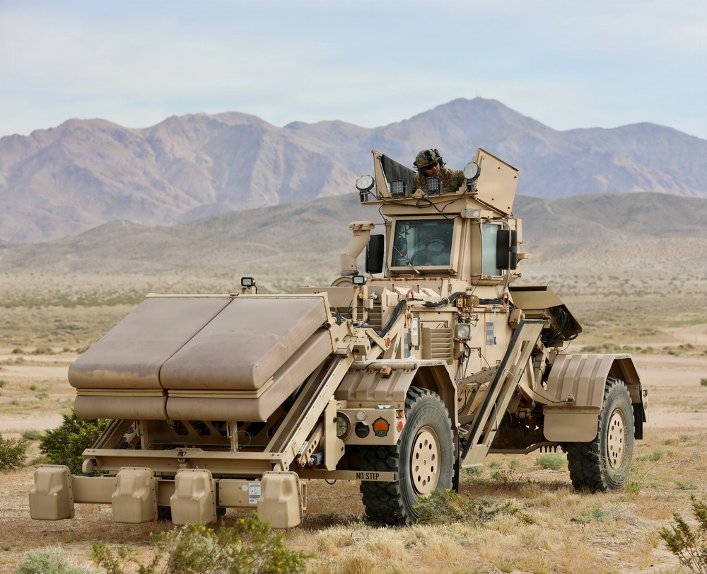 A large tan/desert-camouflaged military vehicle is stationary in an arid desert environment with mountains visible in the background. A soldier in combat gear is visible in the open hatch at the top of the vehicle. The vehicle appears to be a specialized engineering or route-clearance vehicle equipped with front-mounted rollers or mine-clearing attachments.