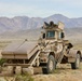 A large tan/desert-camouflaged military vehicle is stationary in an arid desert environment with mountains visible in the background. A soldier in combat gear is visible in the open hatch at the top of the vehicle. The vehicle appears to be a specialized engineering or route-clearance vehicle equipped with front-mounted rollers or mine-clearing attachments.