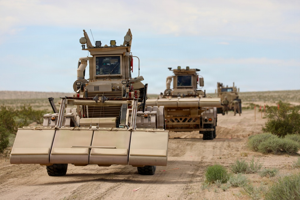 The image shows a convoy of military mine-clearing and route-clearance vehicles traveling along a dirt road in an arid, desert-like environment. The lead vehicle appears to be equipped with a mine-roller or ground-pressure attachment at the front, consistent with IED/mine detection and neutralization equipment. The vehicles are painted in desert tan camouflage, suggesting a military operational or training context.