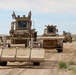 The image shows a convoy of military mine-clearing and route-clearance vehicles traveling along a dirt road in an arid, desert-like environment. The lead vehicle appears to be equipped with a mine-roller or ground-pressure attachment at the front, consistent with IED/mine detection and neutralization equipment. The vehicles are painted in desert tan camouflage, suggesting a military operational or training context.