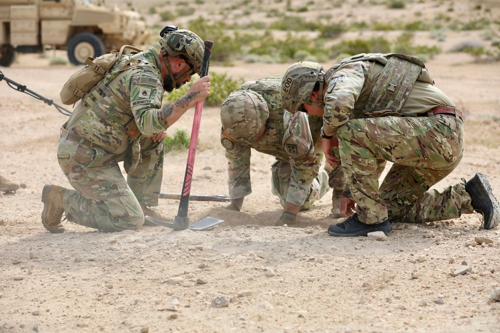 Three military personnel in MultiCam/OCP camouflage uniforms are kneeling and crouching on arid, rocky ground, closely examining a spot in the dirt. One soldier, identified by name tape as 'MERCARTER,' is using a pink-handled shovel to dig or probe the ground. At least one soldier has an 'EOD' (Explosive Ordnance Disposal) label visible on their kit, suggesting this is an IED detection or disposal training exercise.
