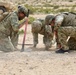 Three military personnel in MultiCam/OCP camouflage uniforms are kneeling and crouching on arid, rocky ground, closely examining a spot in the dirt. One soldier, identified by name tape as 'MERCARTER,' is using a pink-handled shovel to dig or probe the ground. At least one soldier has an 'EOD' (Explosive Ordnance Disposal) label visible on their kit, suggesting this is an IED detection or disposal training exercise.