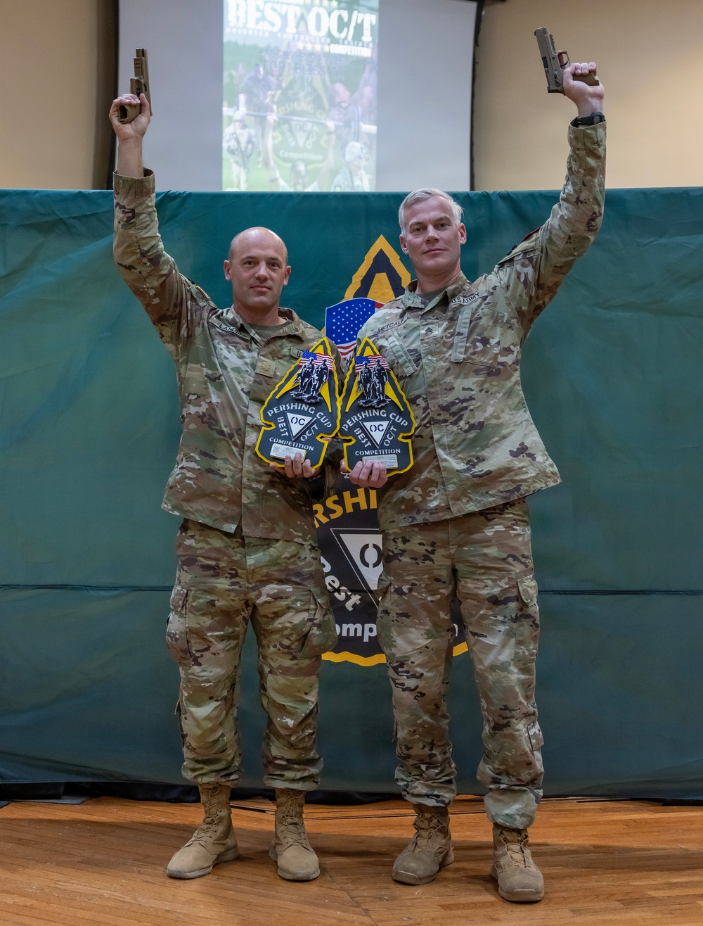 Two U.S. Army soldiers in MultiCam/OCP camouflage uniforms pose for a victory photo, each holding a pistol raised above their head with one hand and a 'Pershing Cup Best OC/T Competition' award plaque in the other. They are standing in front of a backdrop displaying 'Best OC/T' competition branding and a First Army shoulder sleeve insignia. The image appears to document winners of a military training competition.