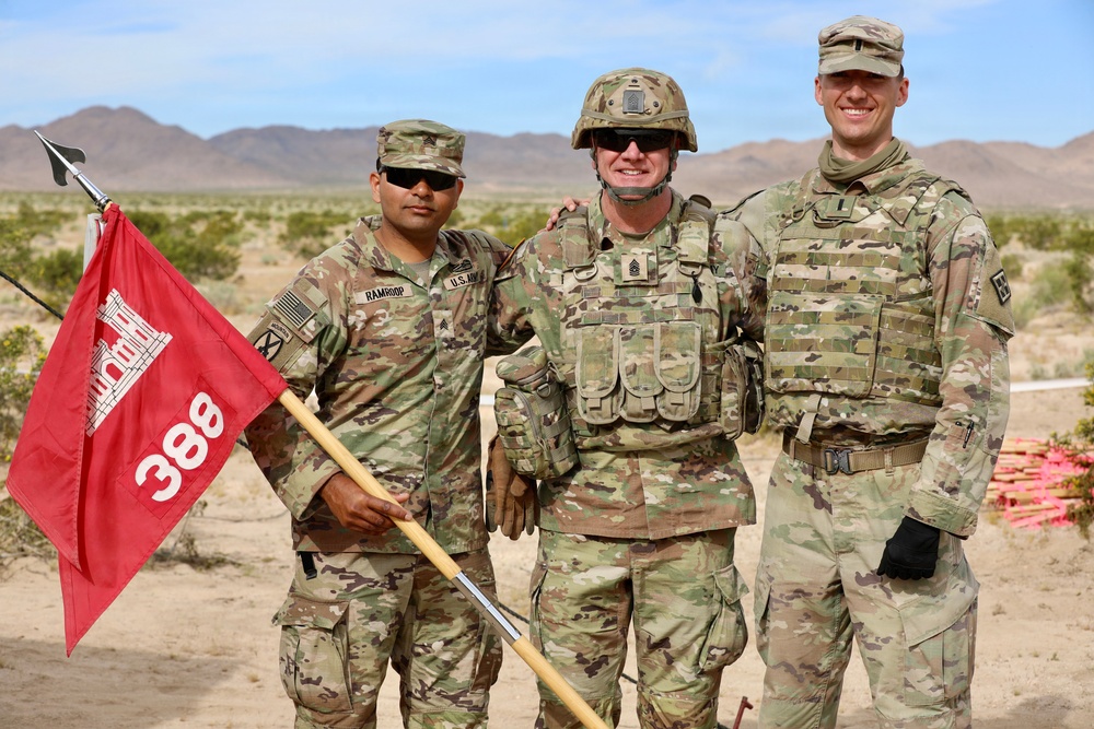 Three U.S. Army soldiers in MultiCam (OCP) camouflage uniforms pose together in a desert training environment. The soldier on the left holds a red guidon flag marked '388' featuring the U.S. Army Corps of Engineers castle insignia. The setting appears to be a field training exercise in an arid, mountainous region.