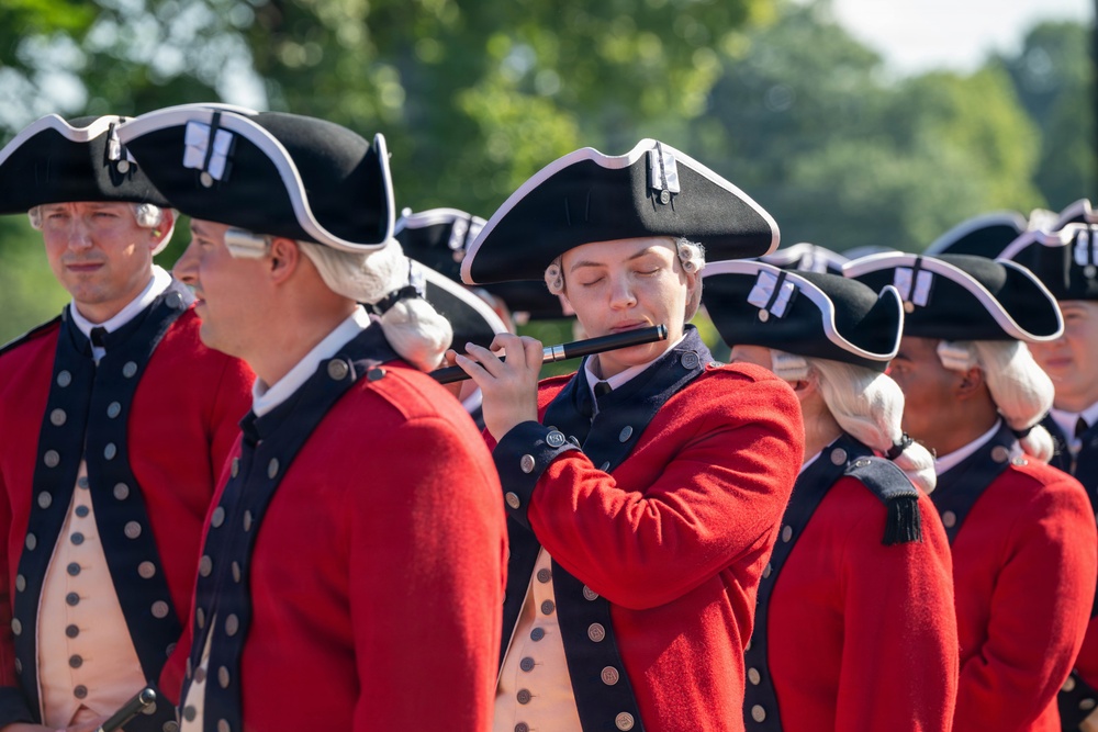 A group of performers dressed in 18th-century American Revolutionary War-era military uniforms are participating in a historical reenactment or ceremonial performance. The central figure is playing a fife (small transverse flute), while others stand in formation around them. All performers wear matching red coats, tricorn hats, white wigs, and tan waistcoats consistent with Continental Army or period military dress.