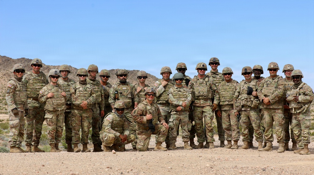 A large group of approximately 20 military personnel in U.S. Army OCP (Operational Camouflage Pattern) uniforms pose for a group photograph in an arid desert environment. The soldiers are wearing combat gear including plate carriers, helmets, and sunglasses, suggesting a training exercise or field operation. The setting appears to be a desert training area, likely in the American Southwest based on the terrain and vegetation.