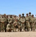 A large group of approximately 20 military personnel in U.S. Army OCP (Operational Camouflage Pattern) uniforms pose for a group photograph in an arid desert environment. The soldiers are wearing combat gear including plate carriers, helmets, and sunglasses, suggesting a training exercise or field operation. The setting appears to be a desert training area, likely in the American Southwest based on the terrain and vegetation.