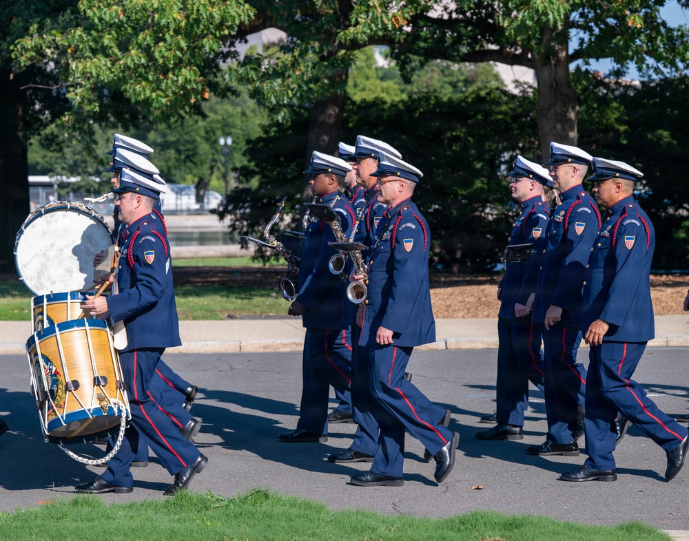 A military marching band is parading in formation along a paved path in what appears to be a park or ceremonial grounds. The musicians are dressed in formal navy blue uniforms with red trim and white-topped caps, carrying instruments including saxophones and a large bass drum. The setting appears to be an outdoor public event, likely in Washington D.C. based on the visible water body and landscape in the background.