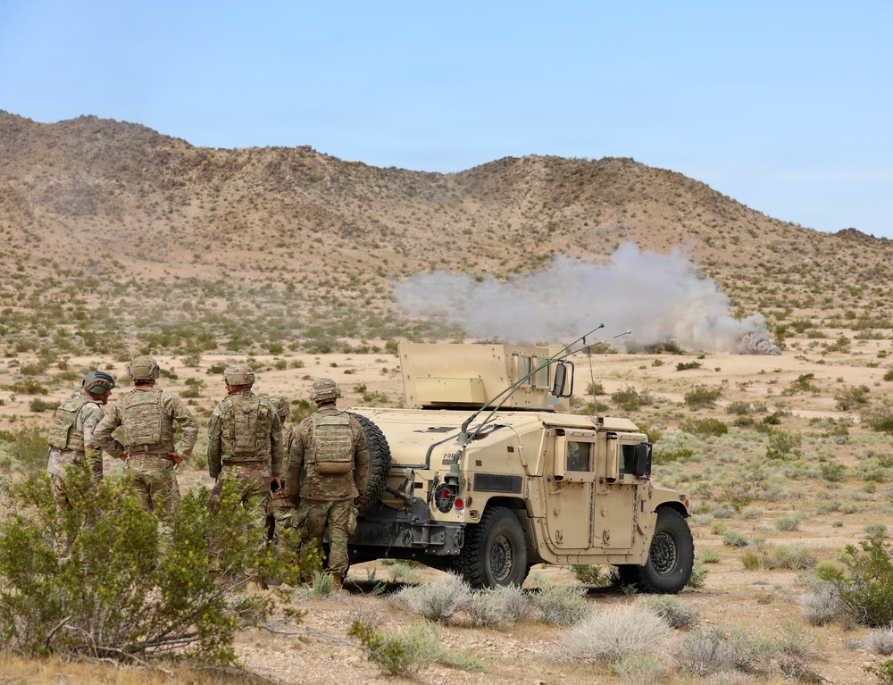 A group of military personnel in camouflage uniforms are observing an explosion or detonation in a desert training environment. They are positioned near a tan-colored HMMWV (Humvee) military vehicle, using it as cover or a vantage point. A visible smoke plume rises in the background near the base of a rocky desert hillside.