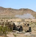 A group of military personnel in camouflage uniforms are observing an explosion or detonation in a desert training environment. They are positioned near a tan-colored HMMWV (Humvee) military vehicle, using it as cover or a vantage point. A visible smoke plume rises in the background near the base of a rocky desert hillside.