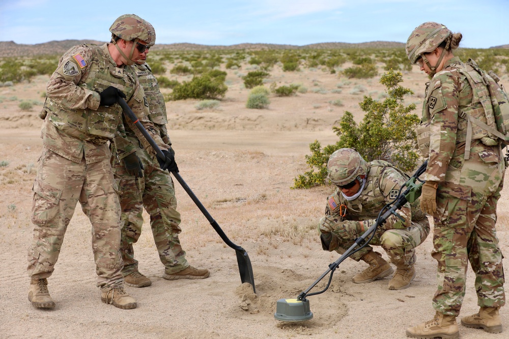 Four U.S. Army soldiers in MultiCam/OCP camouflage uniforms are conducting what appears to be a mine detection and clearance training exercise in a desert environment. One soldier is crouching and using a metal detector while another uses a long-handled tool to probe the ground near a partially exposed simulated landmine or IED training device. The setting appears to be a training range, likely in the Mojave Desert based on the terrain and vegetation.