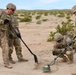 Four U.S. Army soldiers in MultiCam/OCP camouflage uniforms are conducting what appears to be a mine detection and clearance training exercise in a desert environment. One soldier is crouching and using a metal detector while another uses a long-handled tool to probe the ground near a partially exposed simulated landmine or IED training device. The setting appears to be a training range, likely in the Mojave Desert based on the terrain and vegetation.