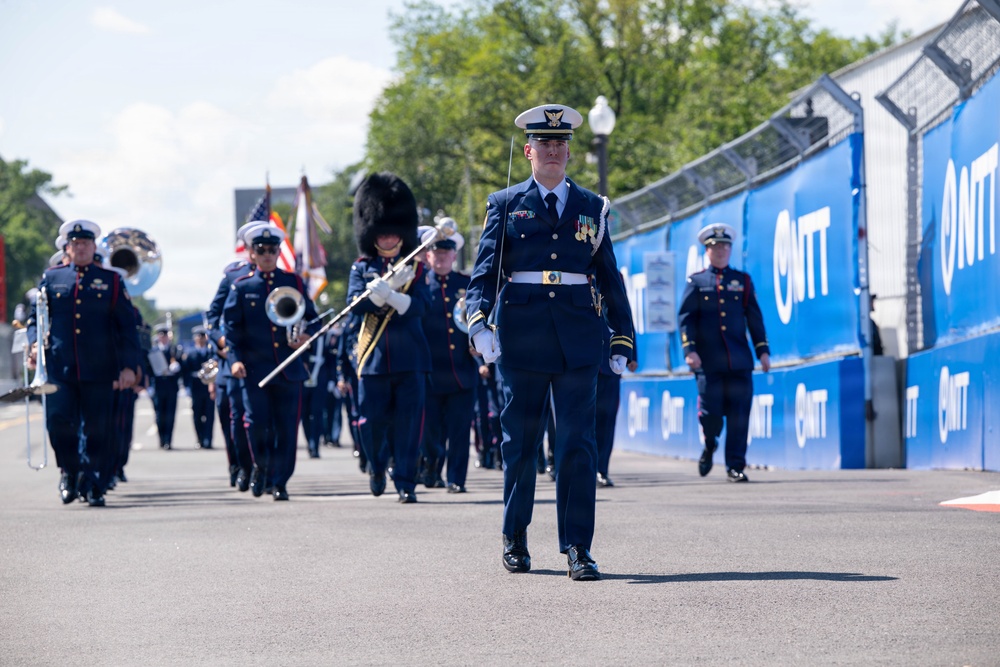 A military marching band parade is taking place along a paved road lined with blue NTT IndyCar Series sponsor banners. The procession is led by an officer in a white-topped dress cap carrying a ceremonial baton, followed by band members in dark navy uniforms playing brass instruments. An American flag and color guard are visible in the formation behind the lead officer.