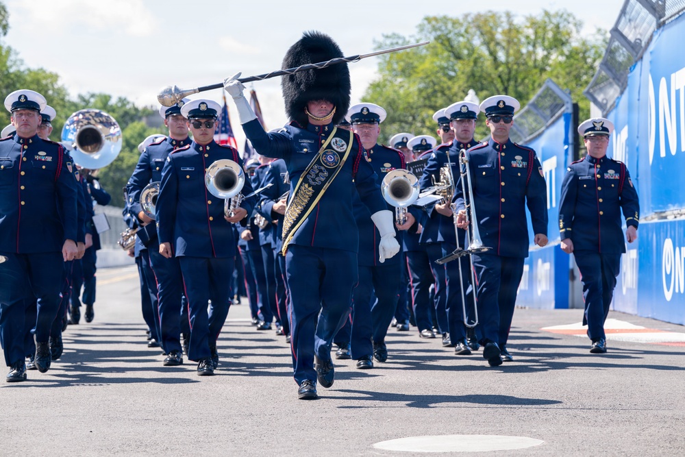 A military marching band is performing in formation along a paved route, led by a drum major wearing a distinctive tall black bearskin hat and carrying a ceremonial mace. The band members are dressed in navy blue uniforms with red trim and white-topped caps, carrying brass instruments including tubas, trombones, and bugles. A blue barrier wall with partial 'NTT' branding is visible on the right side, suggesting this may be at a motorsport venue or similar event.