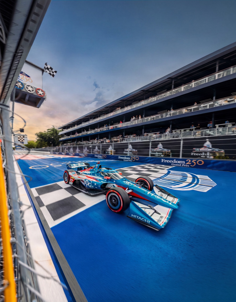 An IndyCar race car crosses the finish line at the Freedom 250 Grand Prix, captured from a low trackside angle. The teal/blue car prominently displays sponsors including PPG and Hitachi, with a patriotic American-themed livery featuring stars and stripes. A checkered flag is visible in the upper left, and spectators fill the multi-level grandstands in the background.