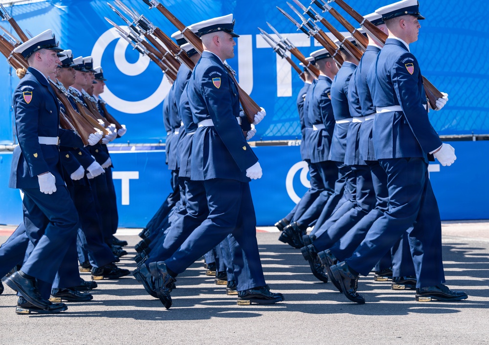 A uniformed military or coast guard honor guard unit is marching in formation during a formal parade or ceremony. The personnel are dressed in matching dark blue dress uniforms with white caps, white gloves, and are carrying rifles with bayonets at a uniform angle. The formation appears highly disciplined and synchronized, taking place in front of a bright blue backdrop with partial white lettering.