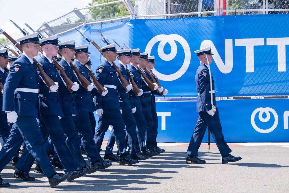A military honor guard unit is marching in formation during a ceremonial parade or event. The personnel are dressed in navy blue dress uniforms with white caps and white gloves, carrying rifles at shoulder arms. The event appears to take place at a motorsport venue, indicated by the NTT sponsorship banners visible in the background.
