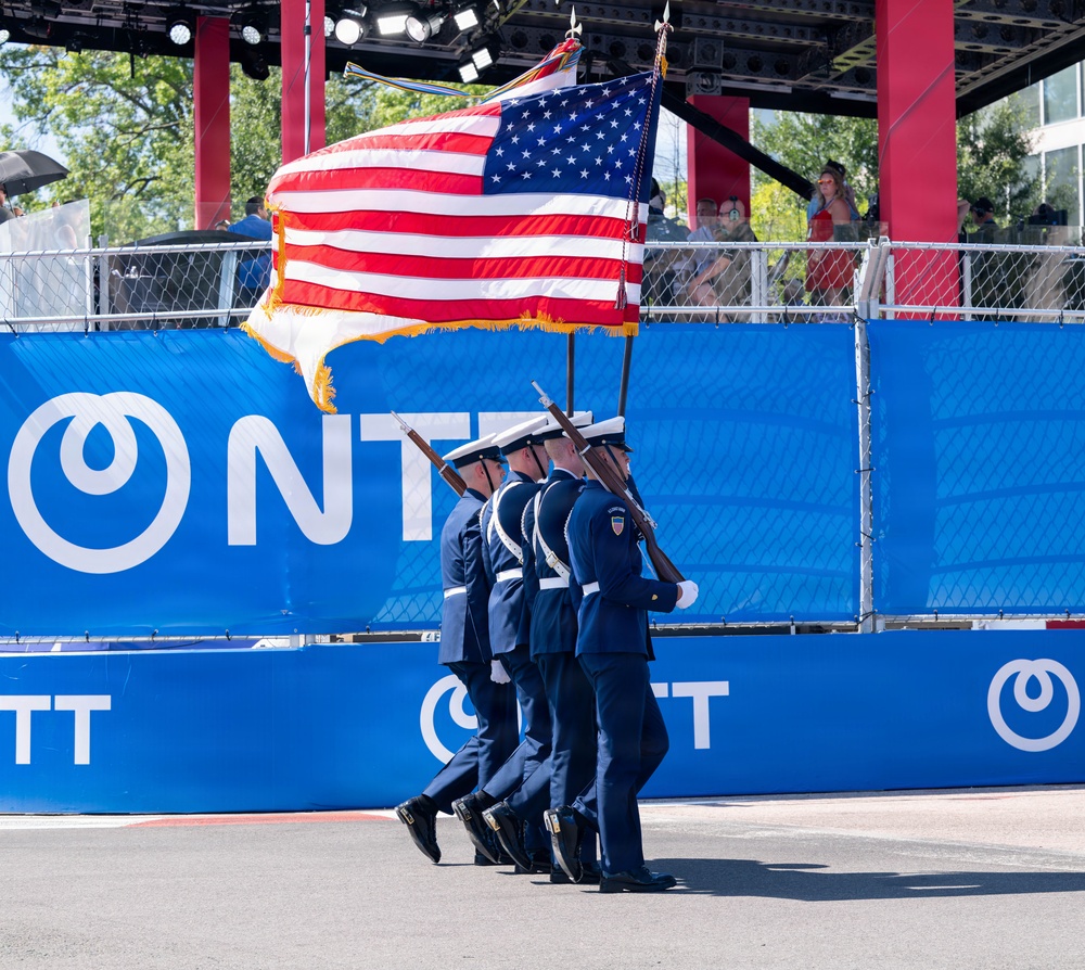 A military color guard in dark blue dress uniforms marches in formation carrying the United States flag and at least one additional flag at what appears to be a motorsport or public event. The personnel are armed with ceremonial rifles and wearing white caps, consistent with a formal color guard presentation. The backdrop features blue sponsor banners with 'NTT' branding, suggesting this may be an IndyCar or similar racing event.