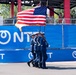 A military color guard in dark blue dress uniforms marches in formation carrying the United States flag and at least one additional flag at what appears to be a motorsport or public event. The personnel are armed with ceremonial rifles and wearing white caps, consistent with a formal color guard presentation. The backdrop features blue sponsor banners with 'NTT' branding, suggesting this may be an IndyCar or similar racing event.