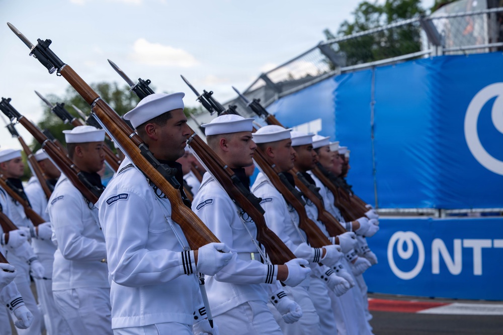 A formation of naval personnel dressed in white uniforms and white sailor caps are marching in a ceremonial parade while carrying rifles with fixed bayonets. The soldiers are wearing white gloves and their uniforms display shoulder patches reading 'CEREMONIAL GUARD'. The event appears to take place near a venue featuring NTT branding on blue barriers in the background.