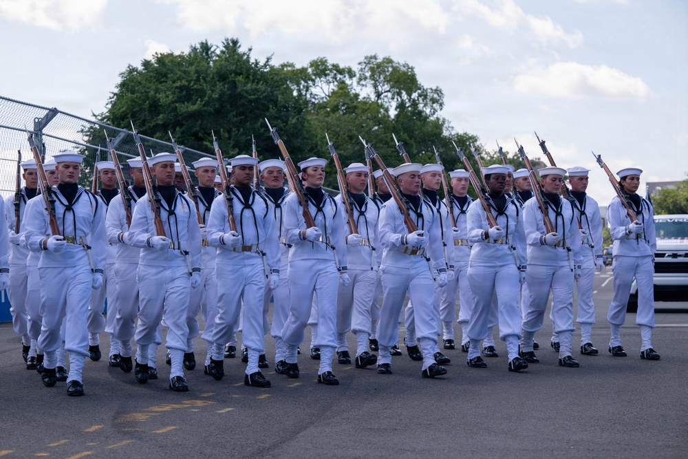 A formation of U.S. Navy personnel in dress white uniforms marches in a ceremonial parade or drill. The sailors are carrying rifles with bayonets at a uniform shoulder-arms position, indicating a formal military ceremony or review. The formation appears well-disciplined and synchronized, suggesting a trained ceremonial unit.