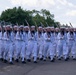 A formation of U.S. Navy personnel in dress white uniforms marches in a ceremonial parade or drill. The sailors are carrying rifles with bayonets at a uniform shoulder-arms position, indicating a formal military ceremony or review. The formation appears well-disciplined and synchronized, suggesting a trained ceremonial unit.