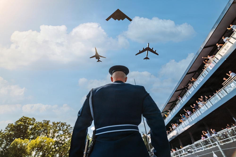 A military officer in dress uniform stands at attention while three aircraft perform a flyover during what appears to be a formal ceremony or graduation event. The aircraft visible include what appears to be a B-2 Spirit stealth bomber, a B-1 Lancer, and a B-52 Stratofortress flying in formation. Spectators in a grandstand to the right are reacting enthusiastically to the aerial display.