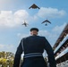 A military officer in dress uniform stands at attention while three aircraft perform a flyover during what appears to be a formal ceremony or graduation event. The aircraft visible include what appears to be a B-2 Spirit stealth bomber, a B-1 Lancer, and a B-52 Stratofortress flying in formation. Spectators in a grandstand to the right are reacting enthusiastically to the aerial display.