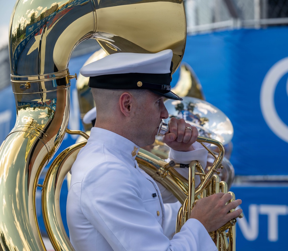 A military musician in white dress uniform is playing a sousaphone (marching tuba) at what appears to be an outdoor ceremonial or parade event. The performer wears a white combination cover (cap) with a black brim, consistent with U.S. Navy or Coast Guard dress whites. A second musician with a similar instrument is partially visible in the background.