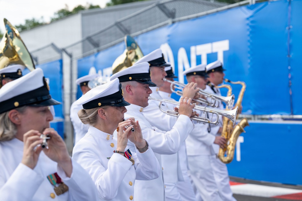 A military band, dressed in white naval dress uniforms with black-brimmed caps, performs outdoors in front of a blue barrier displaying the letters 'NTT'. The musicians are playing a variety of instruments including flutes, trumpets, and saxophones. The setting appears to be a formal or ceremonial outdoor event.