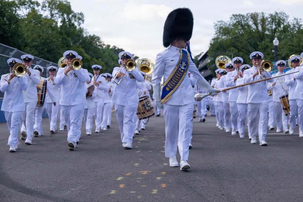 A military marching band is performing in formation on a paved outdoor path, led by a drum major wearing a distinctive tall black bearskin hat and a blue and gold ceremonial sash. All members are dressed in white naval dress uniforms with white peaked caps. The band includes brass instruments such as trombones and tubas, as well as snare drums.
