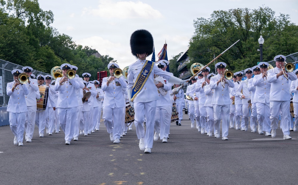 A military marching band in white dress uniforms performs in an outdoor parade or ceremonial event. The drum major leads the formation wearing a distinctive tall black bearskin hat and a blue ceremonial sash, carrying a mace baton. The band consists of numerous musicians playing brass instruments including trombones and trumpets, along with percussion.