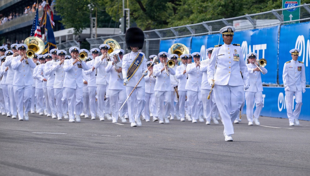 A military marching band in white dress uniforms parades down a street, led by an officer carrying a ceremonial sword and a drum major wearing a tall black bearskin hat. The band members are playing brass instruments including trombones and tubas. Blue sponsor banners line the parade route, and an American flag is visible in the background.