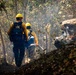 Two wildland firefighters in full protective gear are moving through a smoky forested area toward an active fire. They are wearing standard wildland firefighting equipment including yellow hard hats, fire-resistant Nomex jackets, and backpacks. Flames and smoldering debris are visible to the right side of the frame.