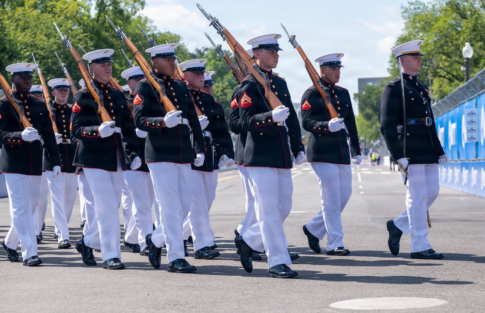 A United States Marine Corps ceremonial drill unit is marching in formation along a paved road, carrying rifles at shoulder arms. The Marines are dressed in their distinctive dress blue uniforms with white trousers, white gloves, and white peaked caps. The setting appears to be a formal parade or public ceremonial event, likely in Washington, D.C., based on the visible blue barriers and tree-lined surroundings.